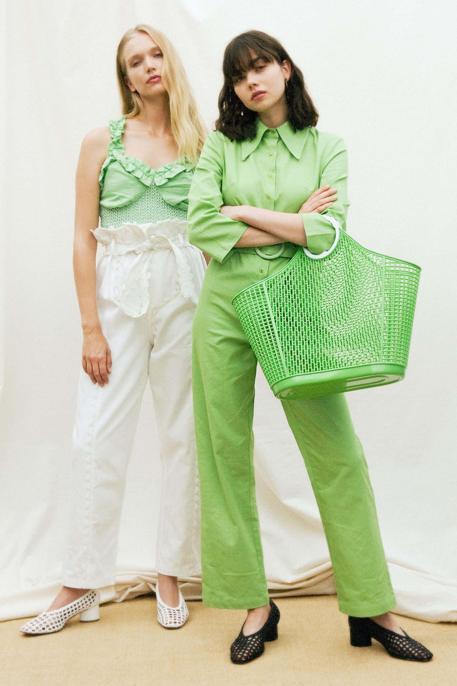 Green Fashion 桌布 壁紙 — 兩位女士在攝影棚自信擺姿勢，一位穿白色褲子和綠色上衣，另一位穿綠色褲子並拿著一個相配的綠色籃子袋。