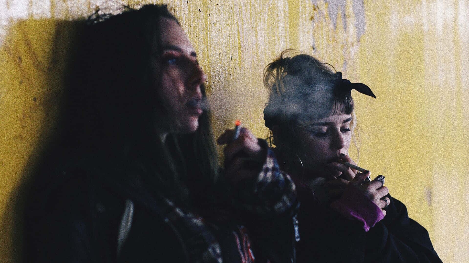 兩位女性靠著黃色牆壁，在陰暗的燈光下抽煙。
