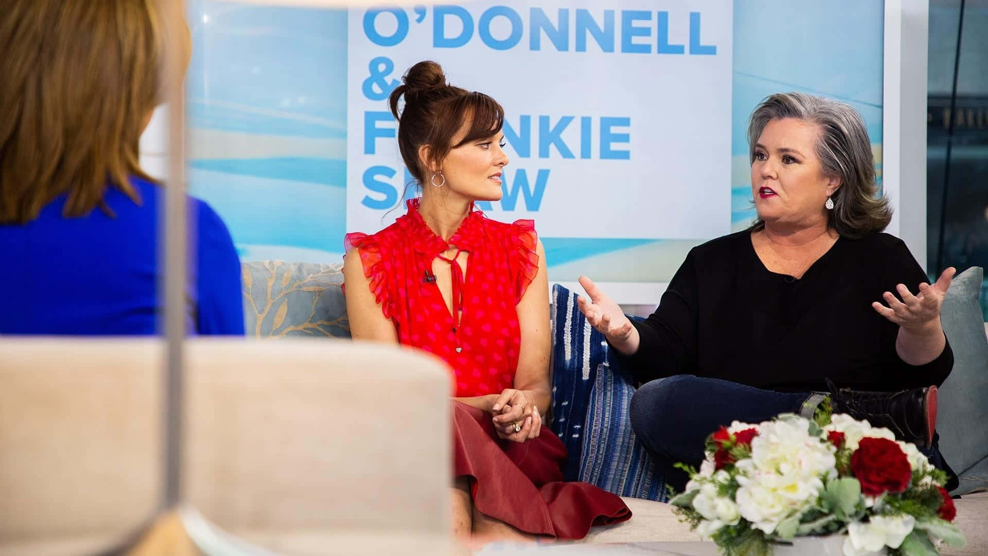 Frankie Shaw 桌布 壁紙 — 兩位女性在日間脫口秀佈景中，以花卉佈置和品牌背景進行熱烈交談。
