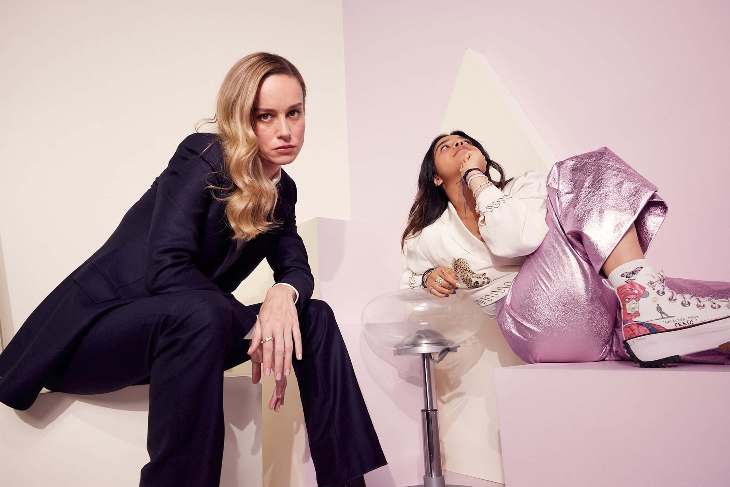 brie larson 桌布 壁紙 — 兩位女性在極簡主義工作室中擺姿勢，一位穿著深色西裝，另一位穿著閃亮的粉紅色家居服搭配圖案運動鞋。