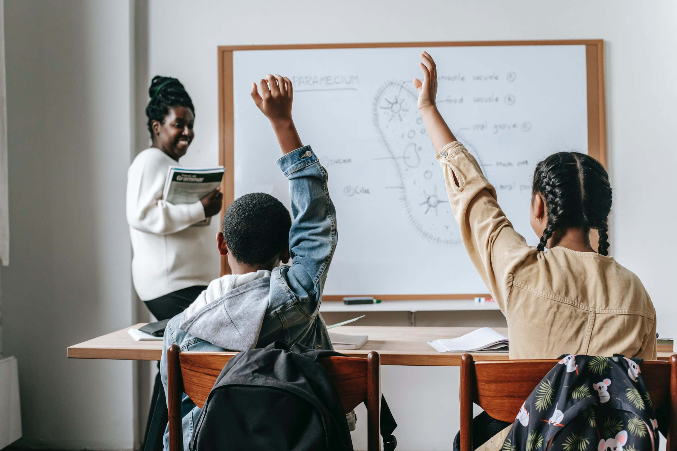 Teacher 桌布 壁紙 — 兩位學生在課室內熱情舉手，一位微笑的老師拿著書本，靠近帶有圖表的白板。