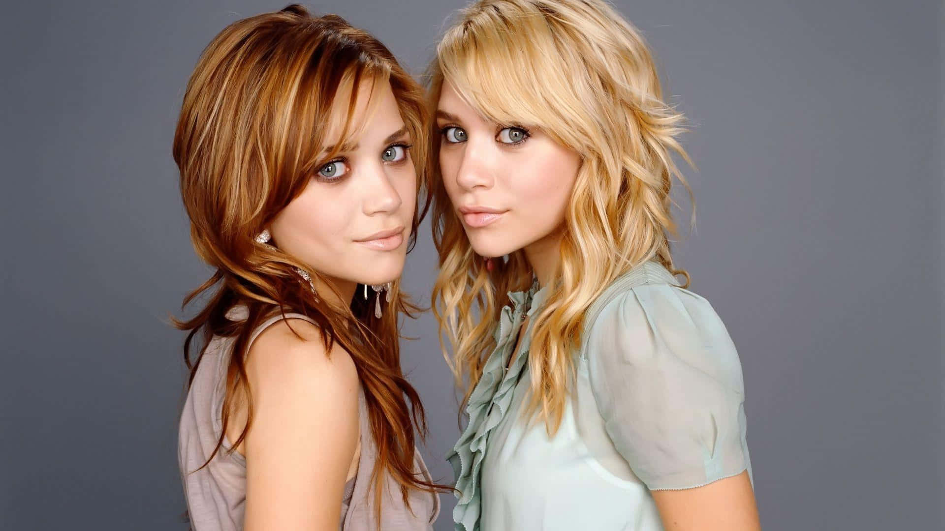 ashley olsen 桌布 壁紙 — 兩位年輕女性，頭髮經過造型，在純灰色背景前緊密地擺姿勢，一位有棕色頭髮，另一位有金色頭髮，兩人都看著相機。
