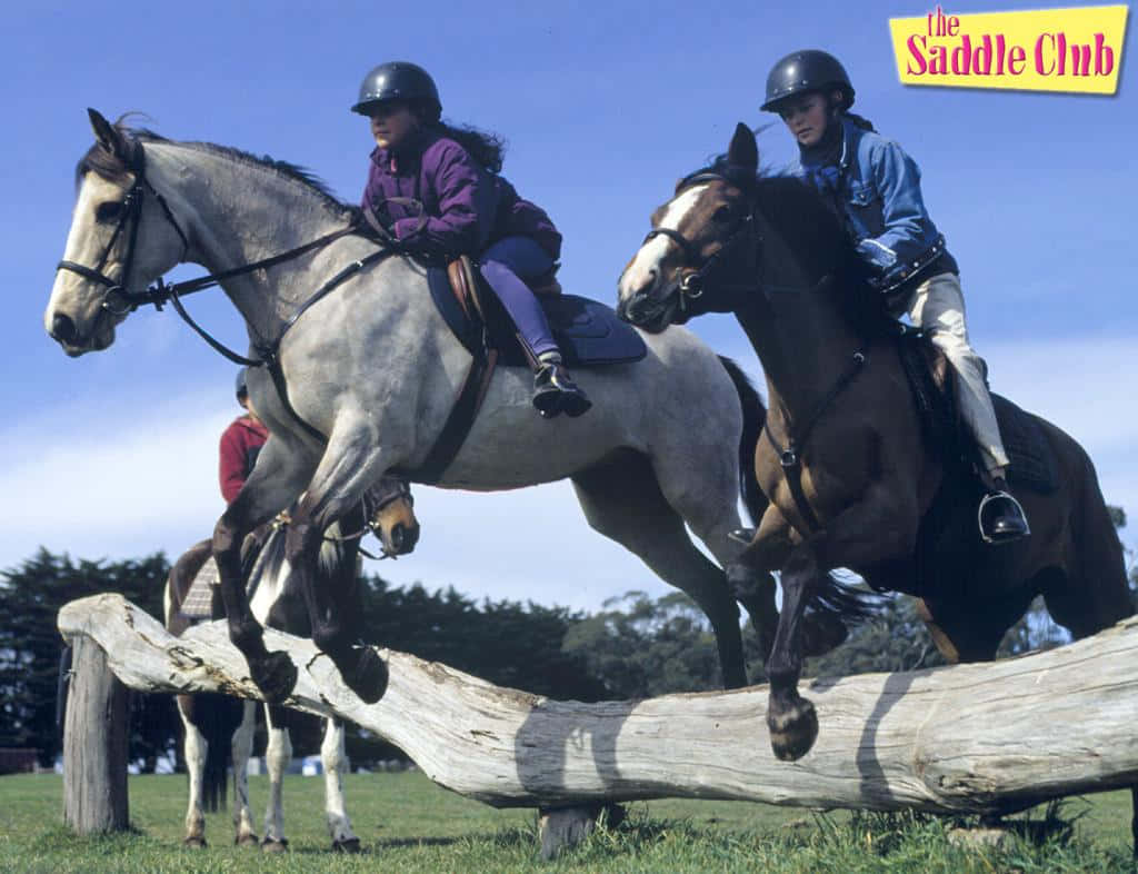 Saddle 桌布 壁紙 — 兩位年輕騎手戴著頭盔，在晴朗的藍天下引導他們的馬匹飛躍一個風化的木柵欄。
