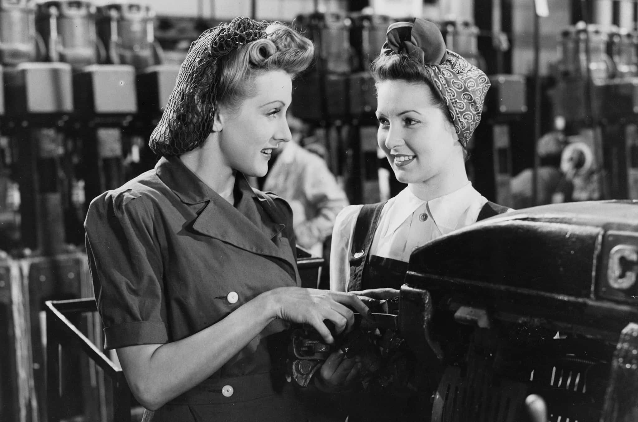 Classic Movie 桌布 壁紙 — 兩位微笑的女性穿著復古工作服，在黑白工廠機器旁分享愉快的時刻。