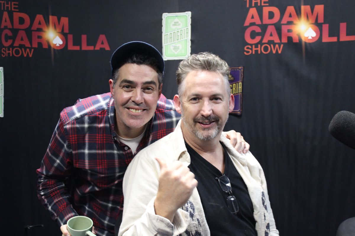  壁紙 — 兩位微笑的男士在 The Adam Carolla Show 中一起擺姿勢，身後有咪高峰和品牌背景。