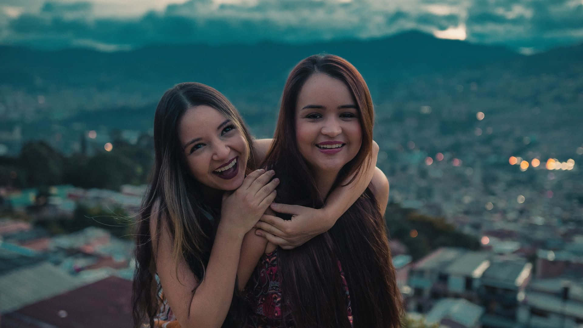 Best Friend 桌布 壁紙 — 兩位快樂的女性在山頂上擁抱，俯瞰黃昏時分廣闊的城市，背景是山脈。