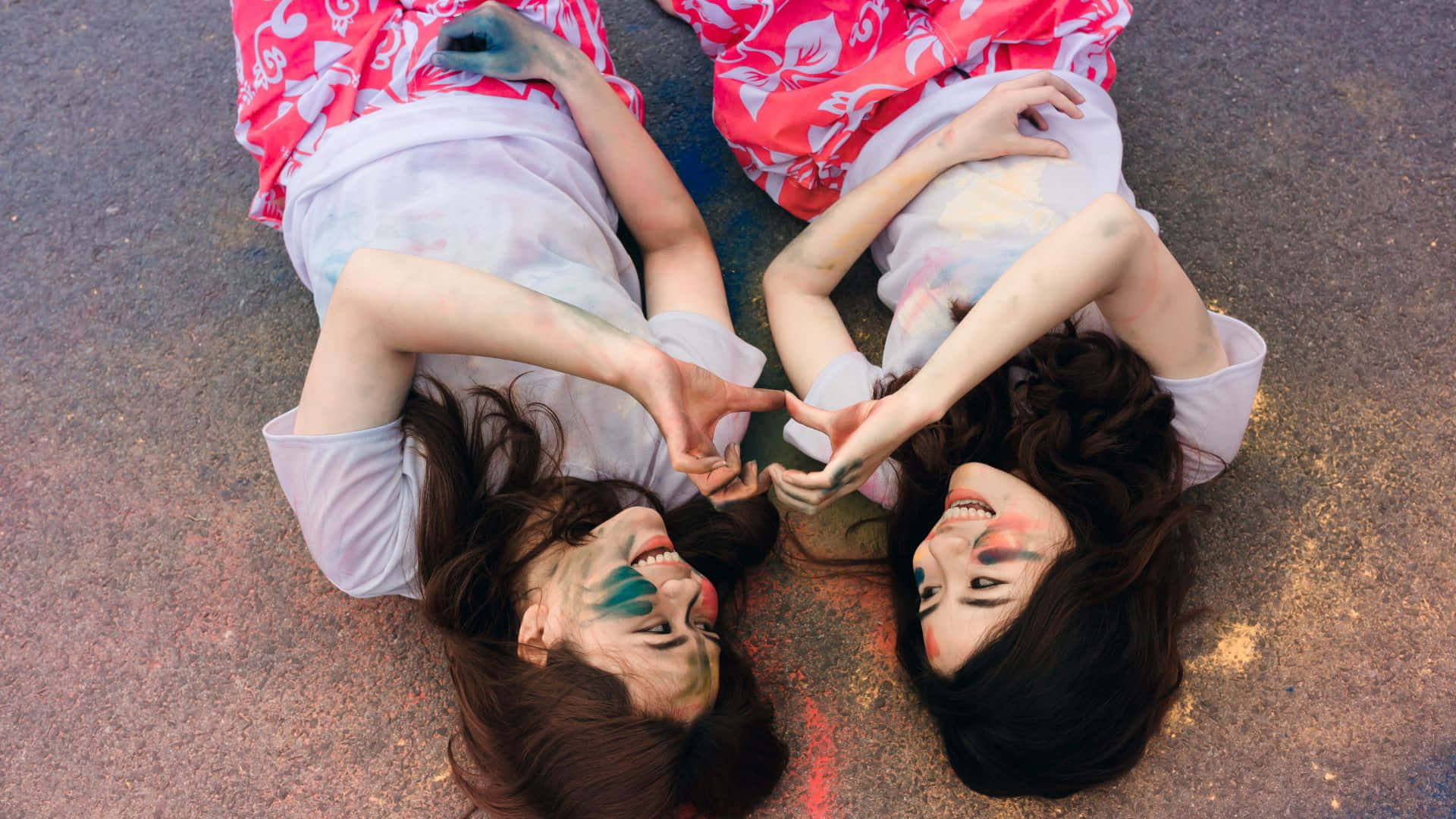 Best Friend 桌布 壁紙 — 兩位快樂的女性臉上塗有彩色顏料，躺在人行道上用手組成一個心形。