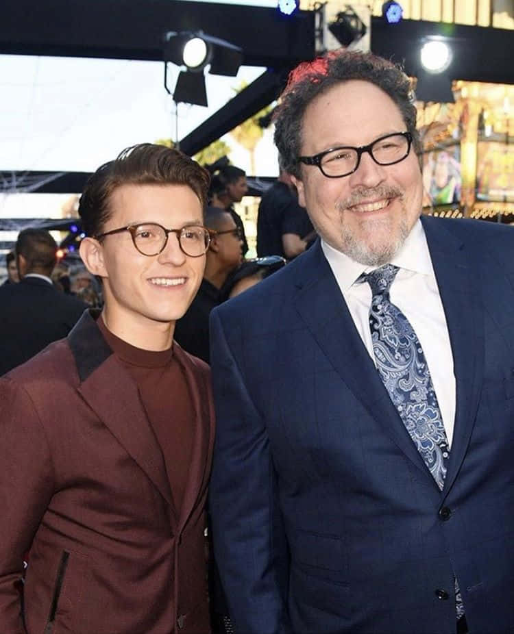 Jon Favreau 桌布 壁紙 — 兩位戴眼鏡的 smiling 男士在舞台燈光下於明亮活動中一起擺姿勢，背後有模糊的人群。