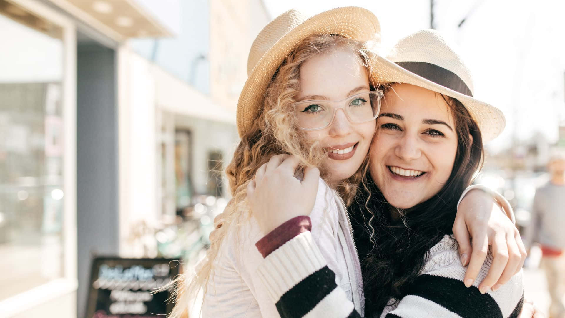 Best Friend 桌布 壁紙 — 兩位戴草帽的年輕微笑女子在明亮的陽光街上熱情擁抱。
