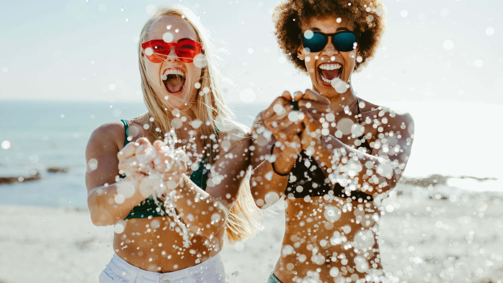 Best Friend 桌布 壁紙 — 兩位戴著太陽眼鏡的快樂女性在陽光明媚的海灘上向鏡頭潑水，閃閃發光的水滴在空中凝固。