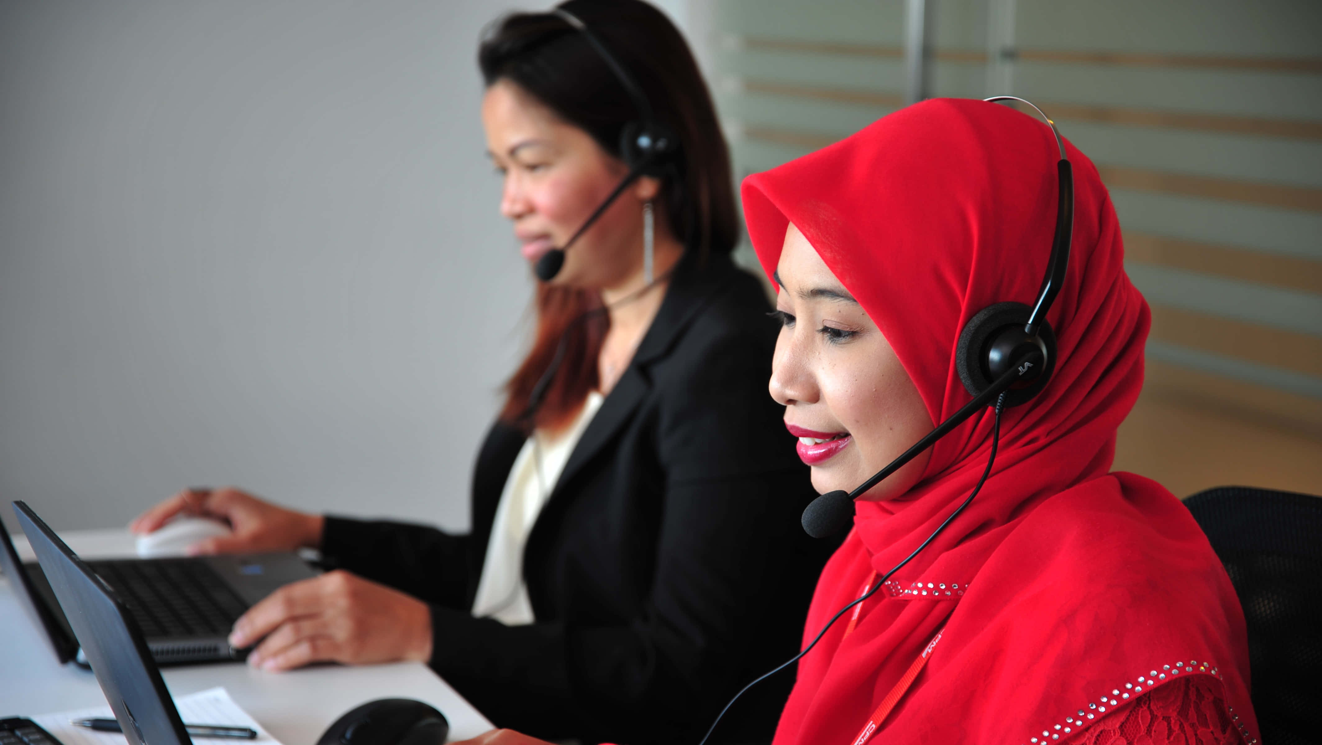 Telemarketing 桌布 壁紙 — 兩位戴著耳機的女性在現代辦公室中，在柔和的燈光和中性色調的牆壁下使用筆記型電腦工作。