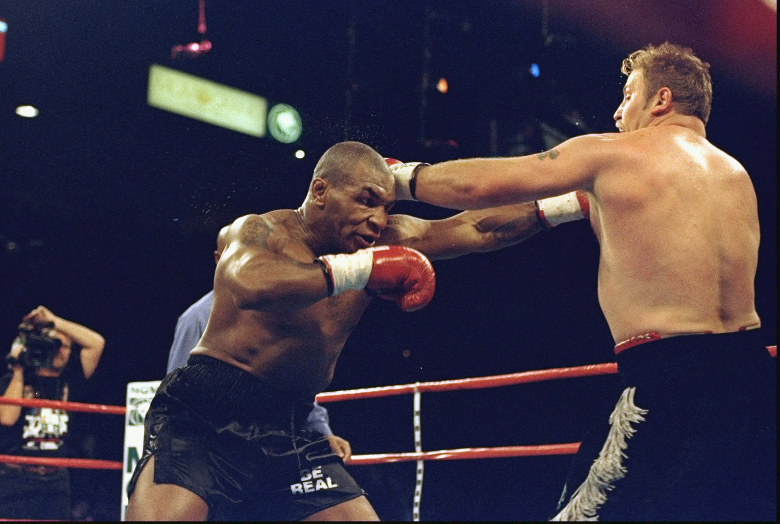 mike tyson 桌布 壁紙 — 兩位拳擊手在擂台上激烈碰撞，汗水飛濺，其中一人在明亮的場館燈光下向對方臉部打出強力一拳。