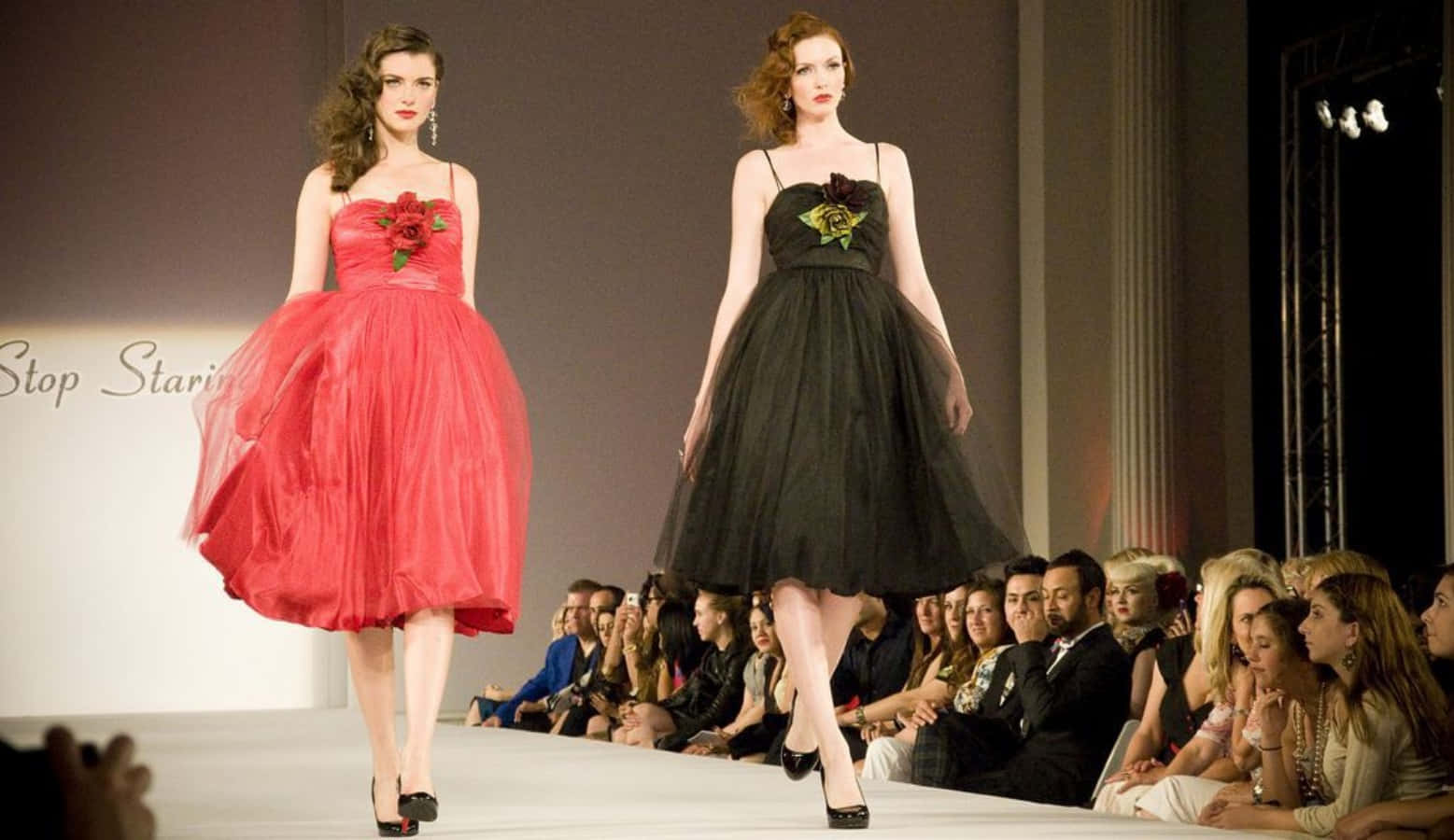 Fashion 桌布 壁紙 — 兩位模特兒穿著寬鬆的連衣裙走秀，一件紅色一件黑色，兩件都飾有花卉裝飾，觀眾專注地觀看。