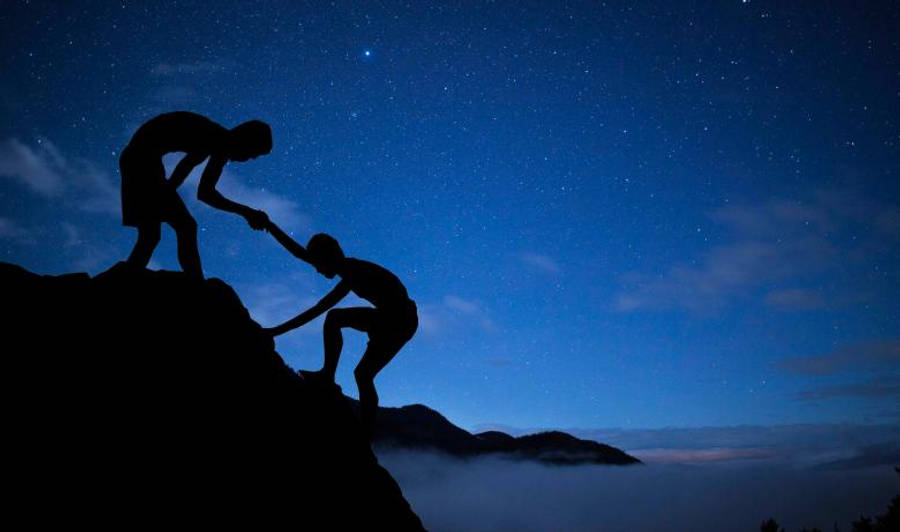 兩位男士的友誼 壁紙 — 剪影人物在星空夜空中互相幫助攀登山峰，下方有雲層。