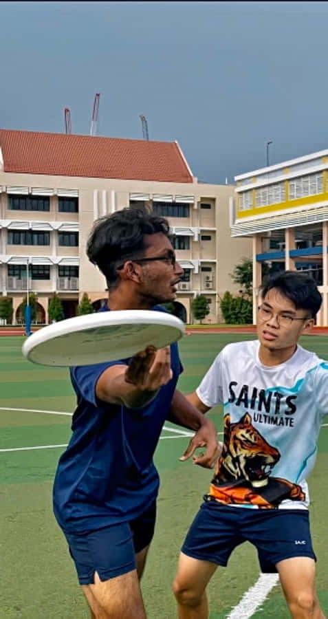 兩位男對手對決 Android Ultimate Frisbee 背景 壁紙 — 兩名年輕男子在綠色運動場上激烈競爭，其中一人將白色飛盤擲向另一人。