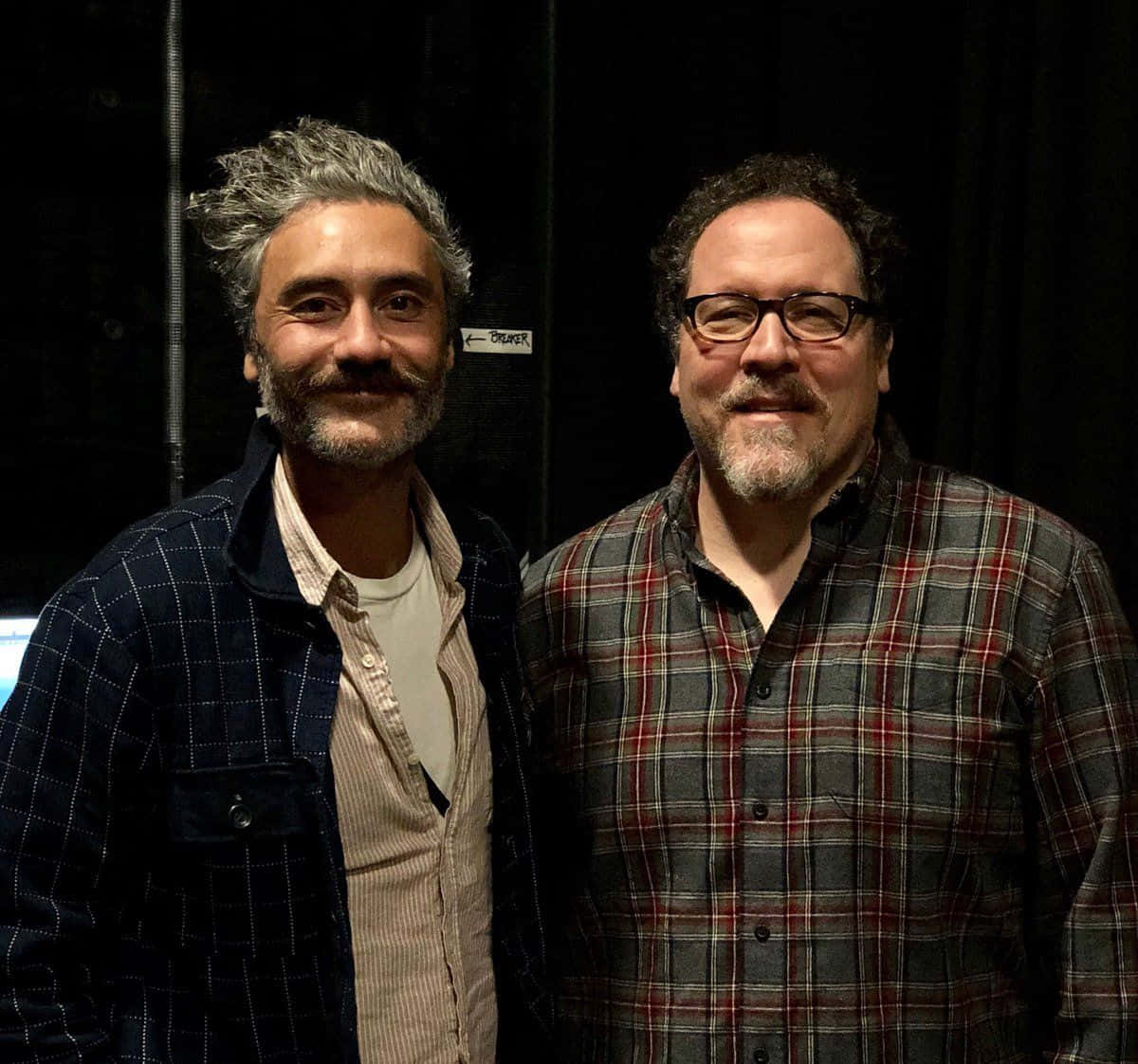Jon Favreau 桌布 壁紙 — 兩位留鬍鬚、身穿格仔恤衫嘅男士喺黑色布幕後台一齊擺甫士微笑。
