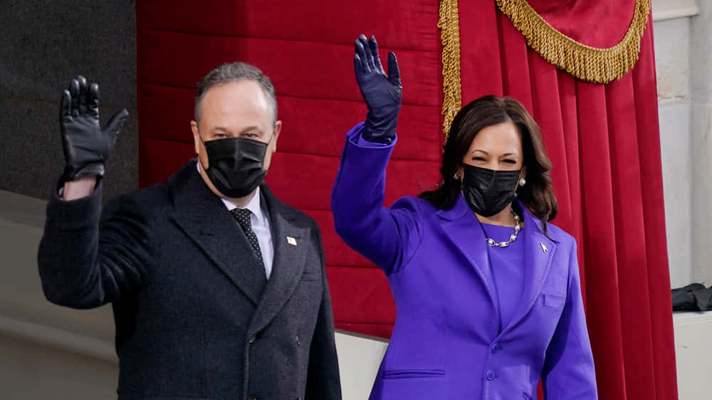Kamala Harris 桌布 壁紙 — 兩位穿著正式服裝的官員戴著黑色口罩，在紅色天鵝絨帷幕背景下向人群揮手。