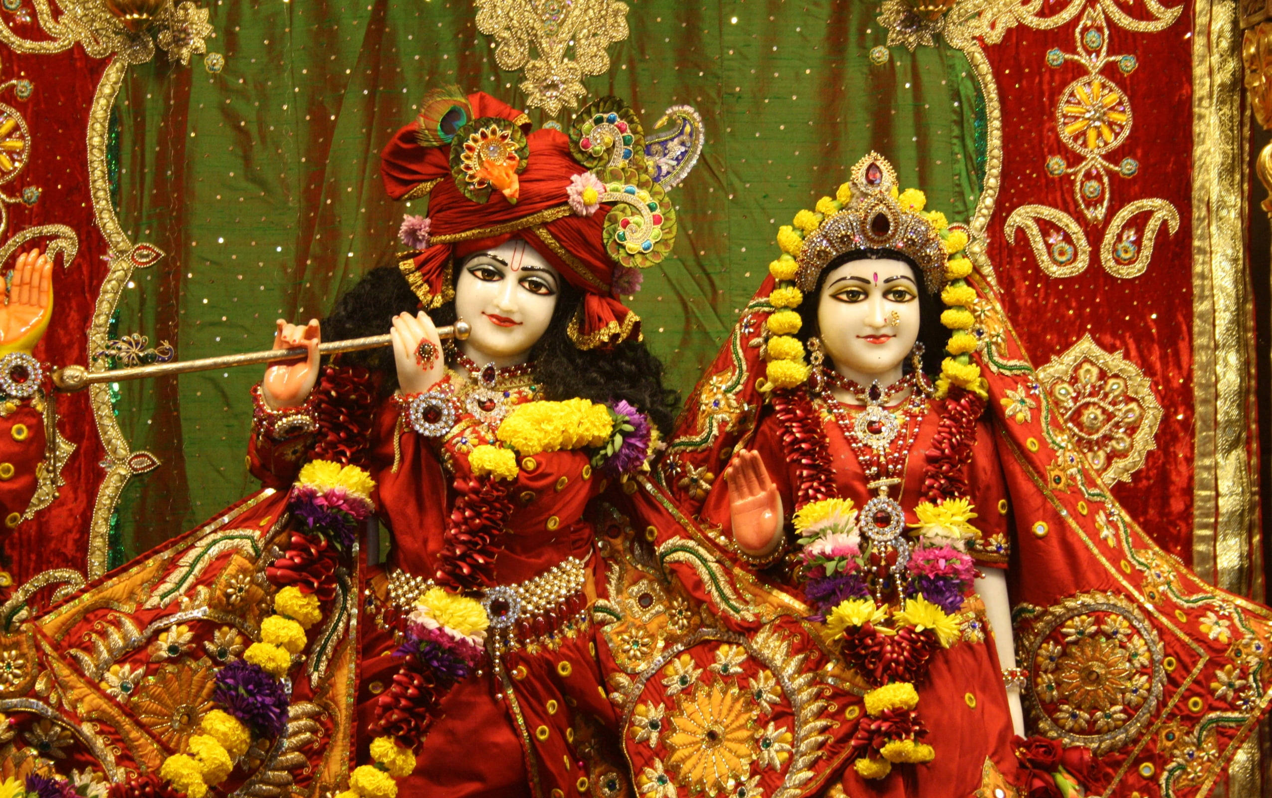 radha krishna HD 桌布 壁紙 — 兩位穿著華麗的印度教神祇，飾以紅、金、黃色花環，站在繡工精緻的綠色和紅色背景前。