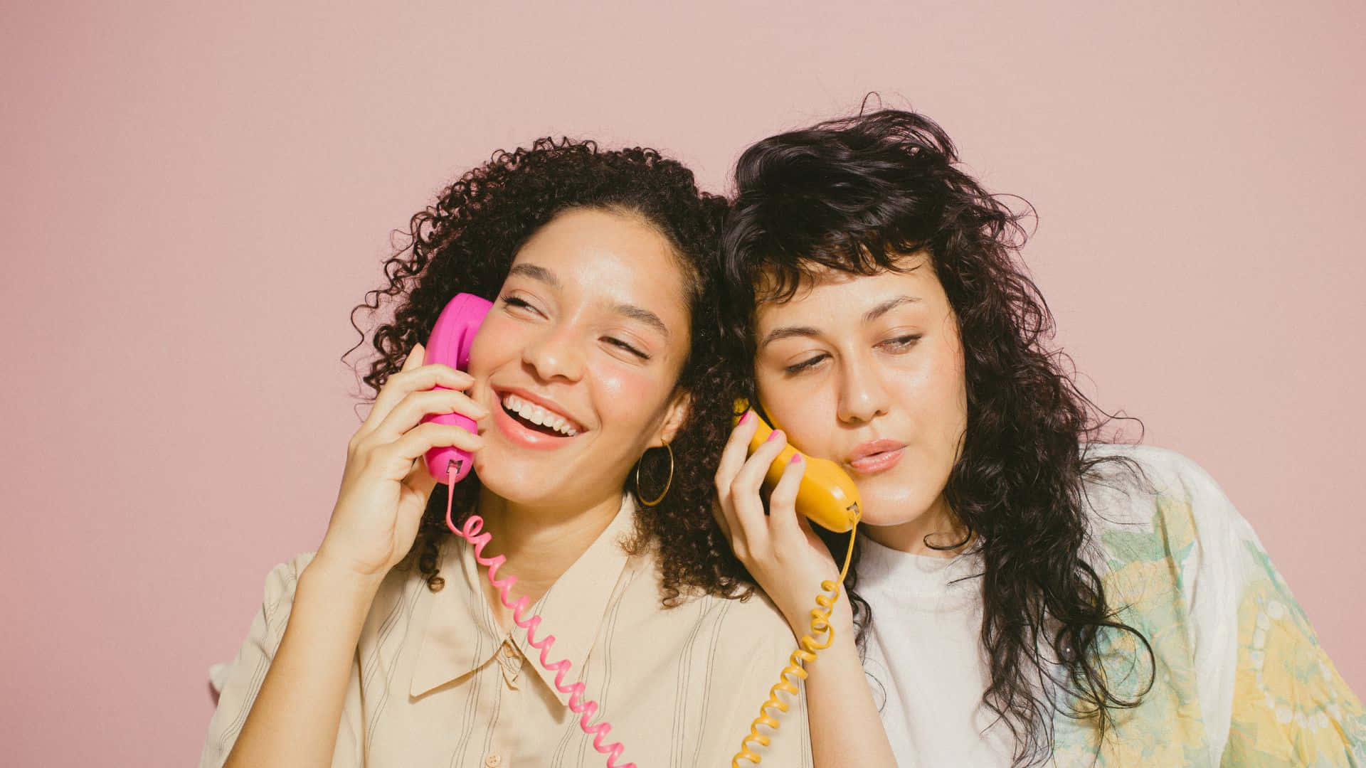 Best Friend 桌布 壁紙 — 兩位笑容滿面、捲髮的女性在柔和的粉紅色背景前，一邊用色彩繽紛的復古轉盤電話交談，一邊大笑。