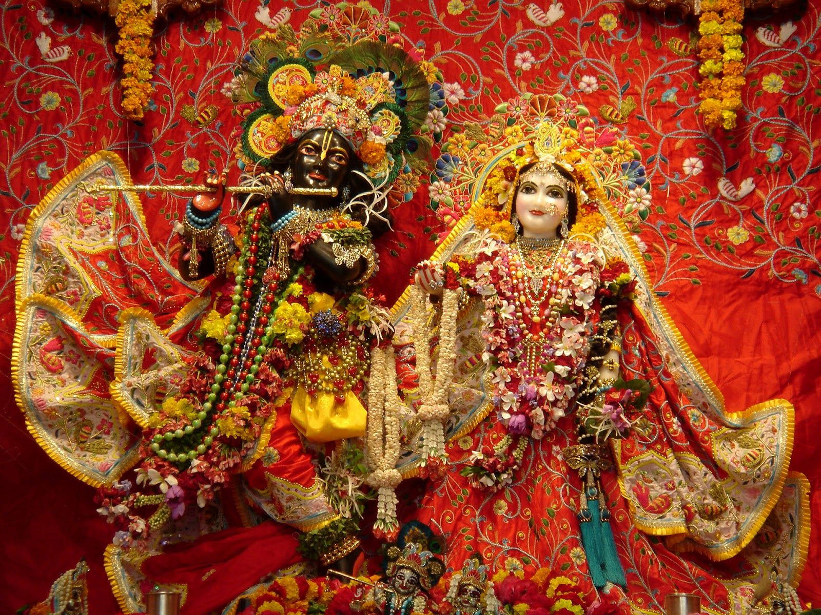Krishna Flute 桌布 壁紙 — 兩位精心裝飾的印度教神祇並排坐在華麗的紅色刺繡背景前，周圍環繞著鮮豔的花環和精緻的珠寶。