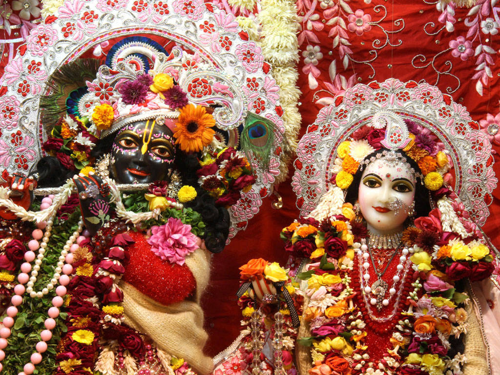 Hindu God 桌布 壁紙 — 兩位裝飾華麗的印度教神祇，披掛著鮮豔的花環和精緻的珠寶，背景是豐富的紅色刺繡。