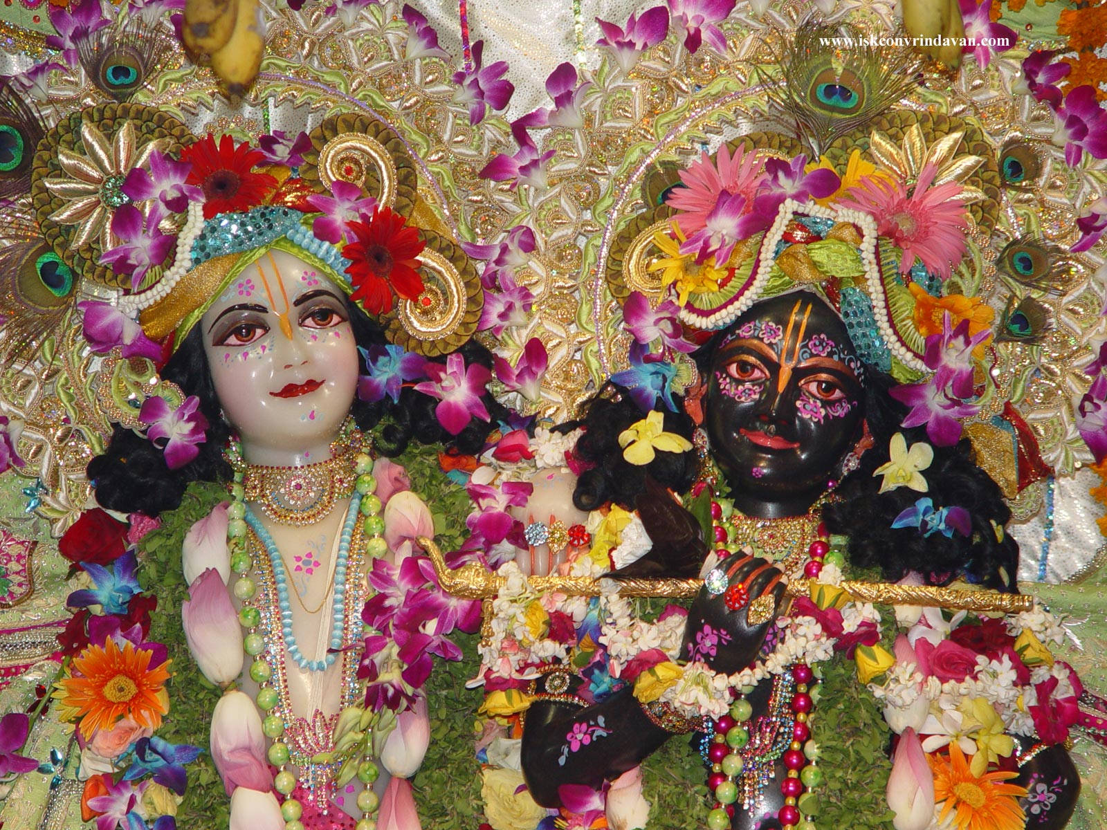 Hindu Temple 桌布 壁紙 — 兩位裝飾華麗的印度教神祇，飾有鮮豔的花朵、孔雀羽毛和金飾，坐在茂盛的花卉佈置中。