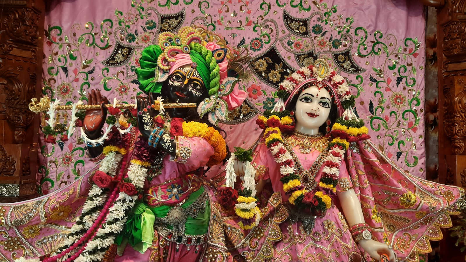 Iskcon 桌布 壁紙 — 兩位裝飾華麗的印度教神祇並排坐在華麗刺繡的粉紅色背景前，飾有鮮花和珠寶。
