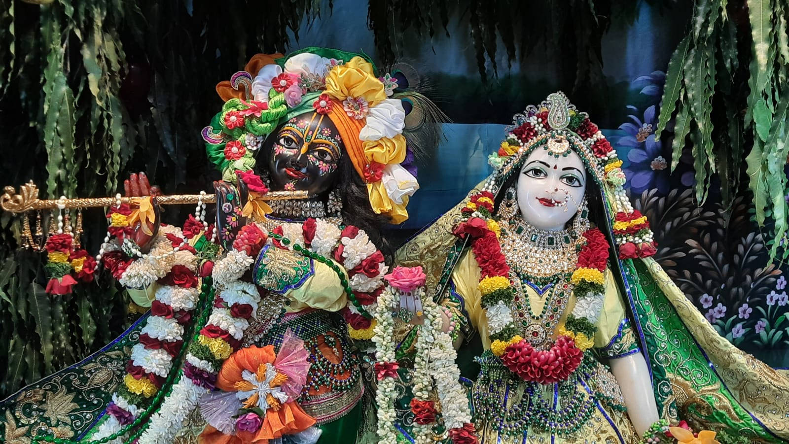 Hindu God 桌布 壁紙 — 兩位裝飾華麗的印度教神祇克里希納和拉達坐在一起，披著鮮豔的花環，背景是茂密的叢林。