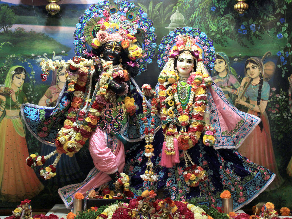 Iskcon 桌布 壁紙 — 兩位裝飾華麗的印度教神祇端坐在色彩繽紛的花卉供品和繪製的神話場景之中，氣勢非凡。