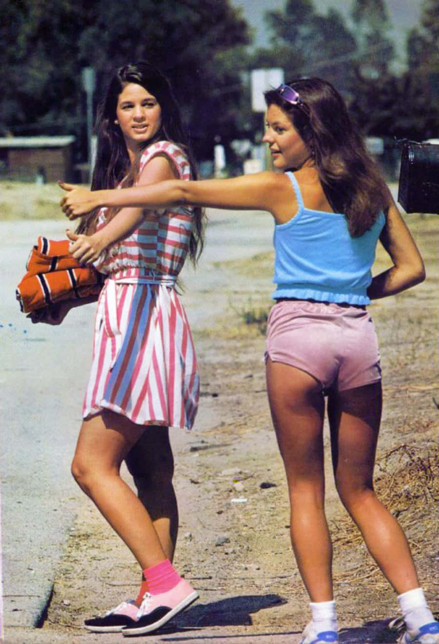 70年代gif 桌布 壁紙 — 兩位身穿1970年代復古服裝的年輕女性在陽光明媚的路邊，以樹木和遠處的建築物為背景，俏皮地擺姿勢。