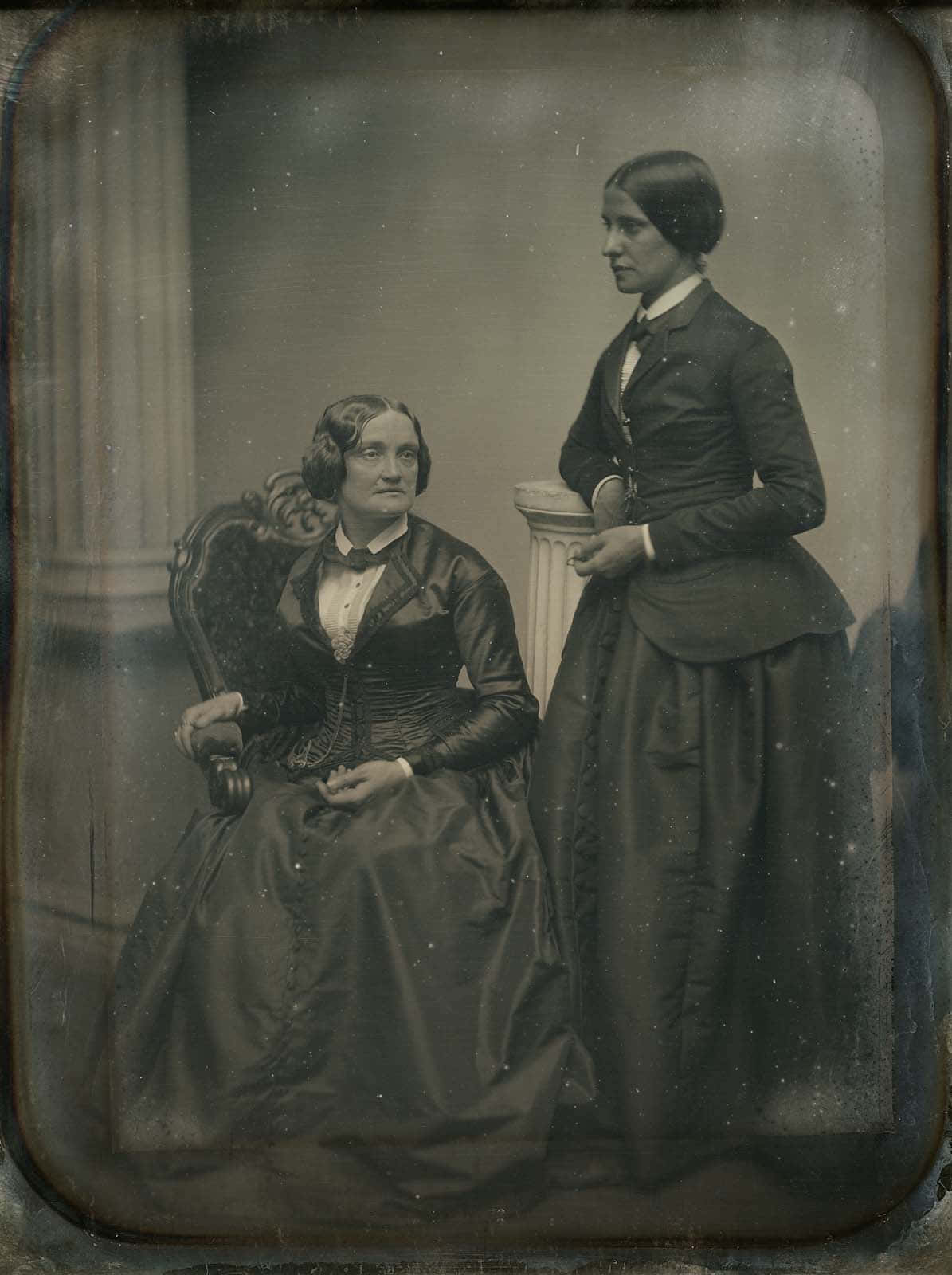 Victorian 桌布 壁紙 — 兩位身穿19世紀正式服裝的女性，一位坐著一位站著，在復古錫版照片中莊嚴地擺姿勢。