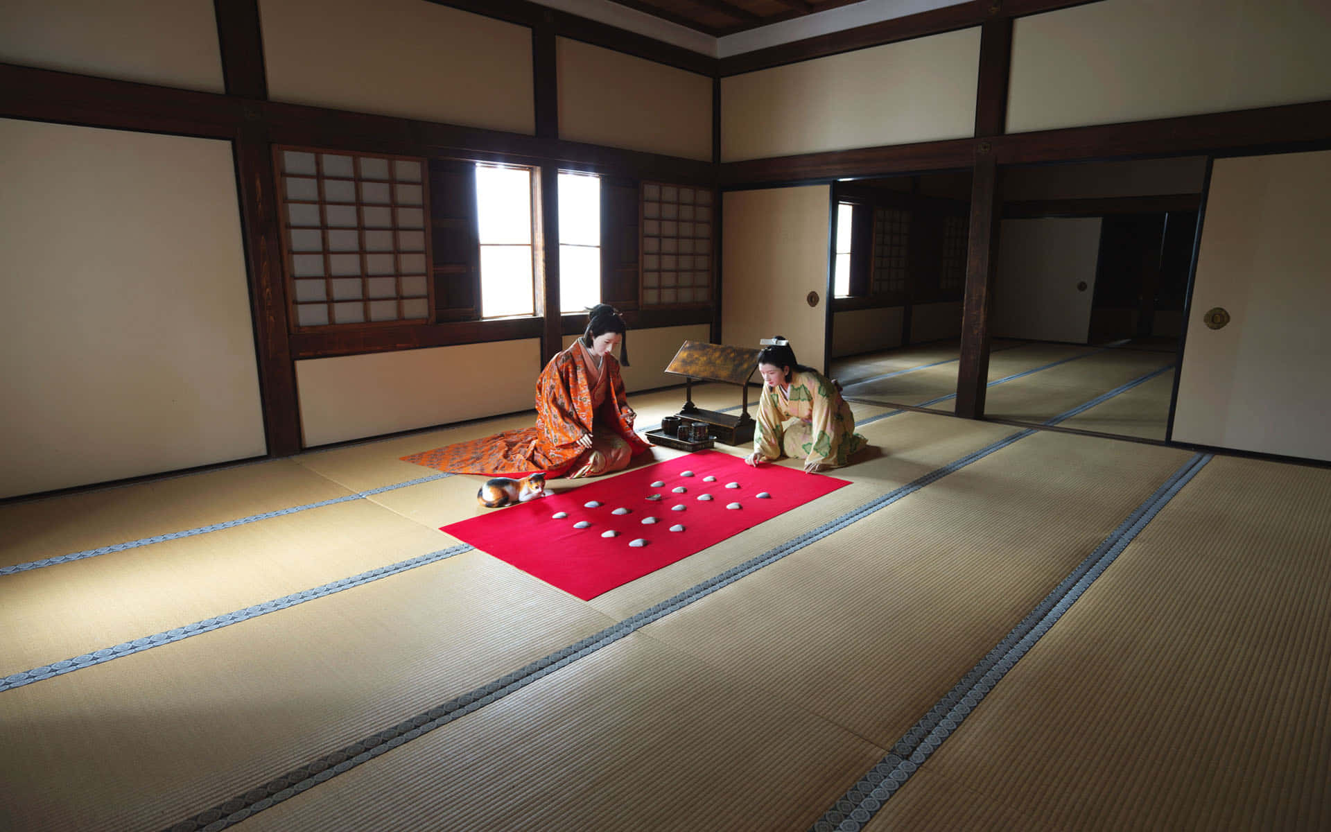 Feudal Japan 桌布 壁紙 — 兩位身穿傳統和服的女性在寧靜的榻榻米房間中，於紅色墊子上對弈圍棋，房間沐浴在柔和自然光中。