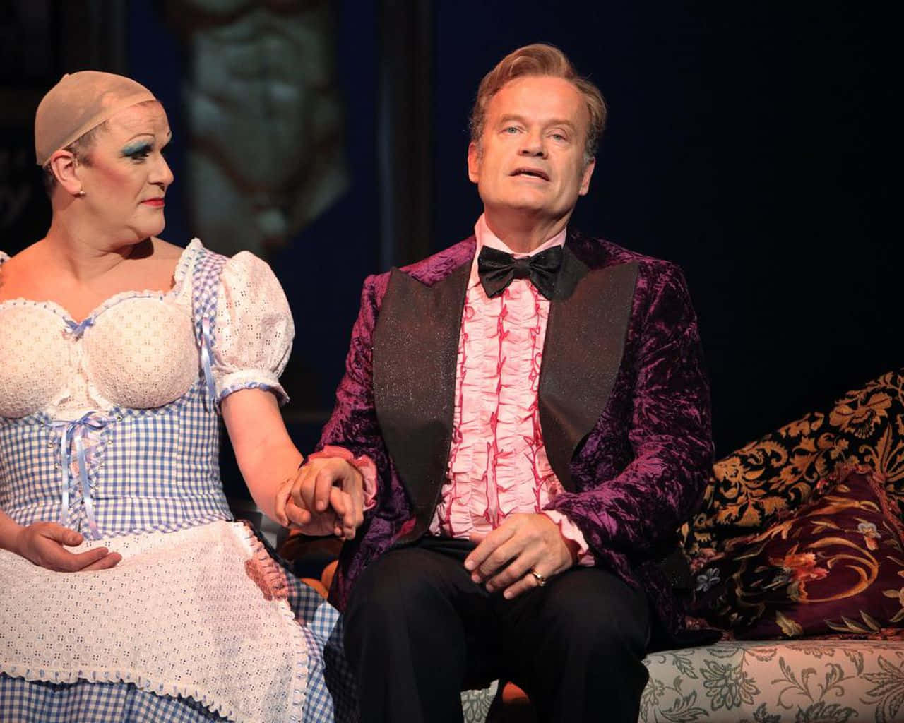 Kelsey Grammer 桌布 壁紙 — 兩位身穿戲劇服裝的演員在裝飾華麗的枕頭和黑暗背景的舞台上牽手。