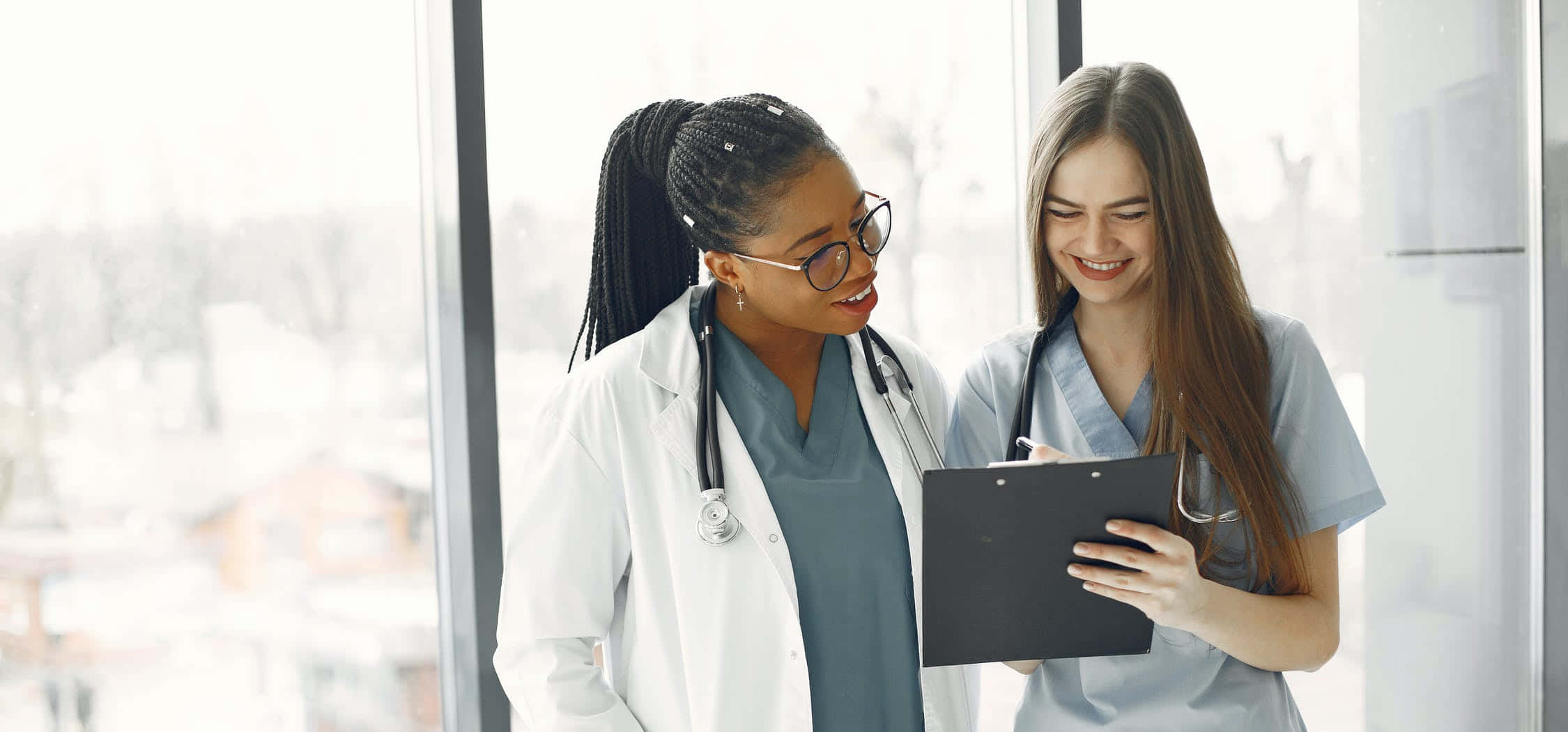 Nursing 桌布 壁紙 — 兩位身穿手術衣和實驗室外套的 smiling 女性醫療專業人員在明亮的窗邊一起檢視記事板。