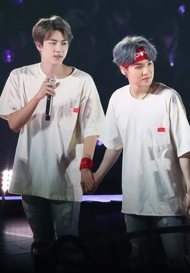 Suga 桌布 壁紙 — 兩位身穿白恤衫的男表演者在昏暗的舞台上，在紫色聚光燈下牽手。
