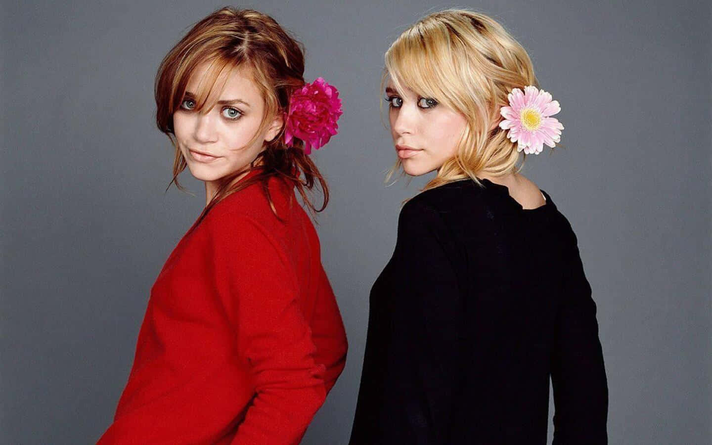 Mary Kate Olsen 桌布 壁紙 — 兩位身穿紅色和黑色上衣的女性，頭髮上插著花朵，在灰色背景前轉向鏡頭。