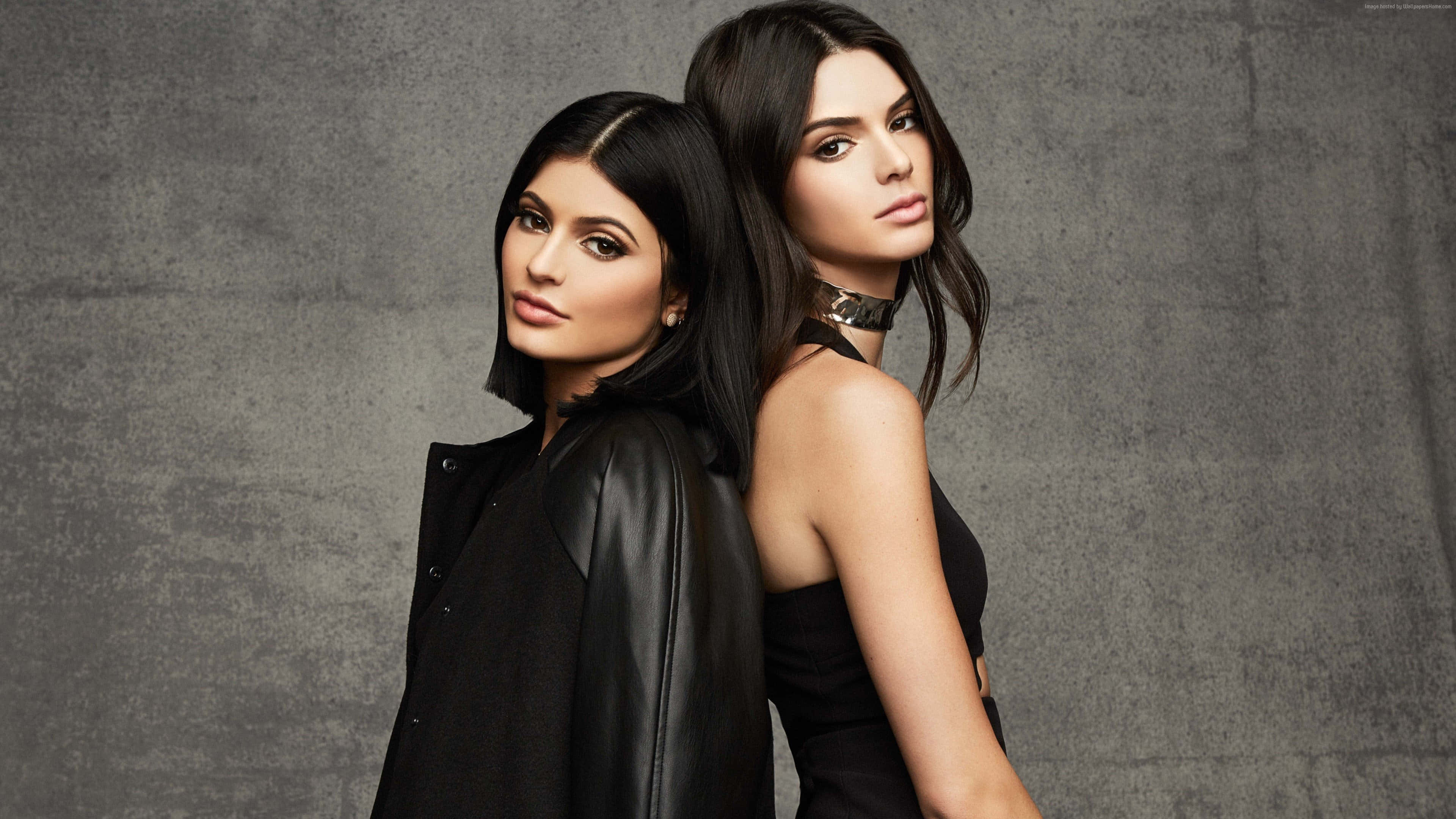 Kylie Jenner 桌布 壁紙 — 兩位身穿黑色服裝的女性背對背，在帶有紋理的灰色工作室牆前擺出自信的表情。