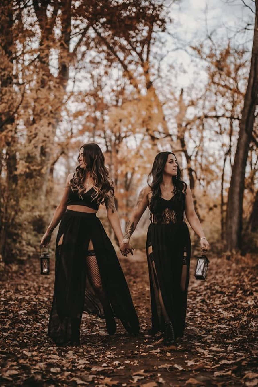 Witch Costumes 桌布 壁紙 — 兩位身穿黑色蕾絲連衣裙的女性手牽手，提著燈籠走過金色的秋林。