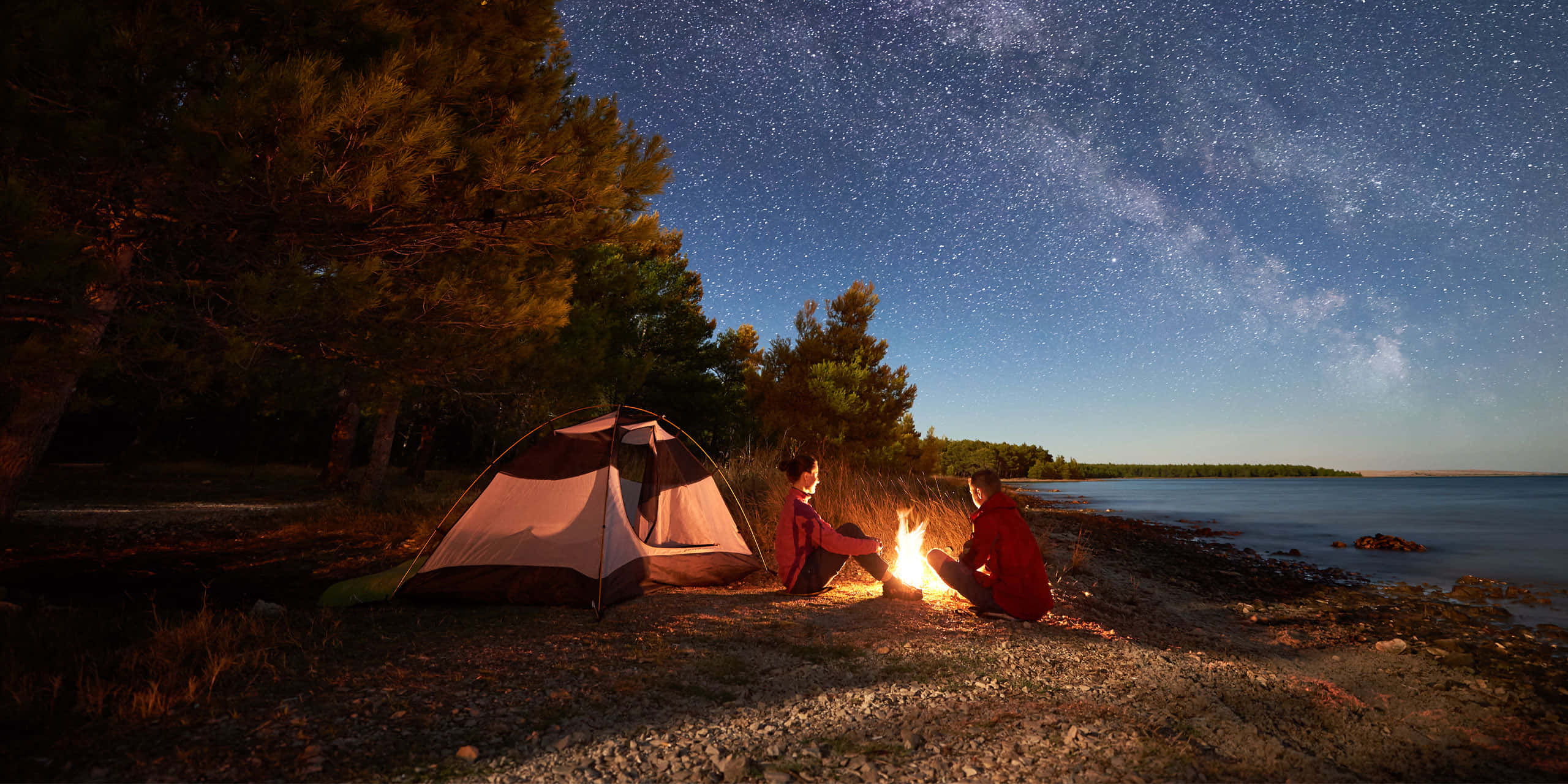 Beach Camping 桌布 壁紙 — 兩位露營者坐在發光的營火旁，星空下銀河拱跨平靜的湖泊和松樹。