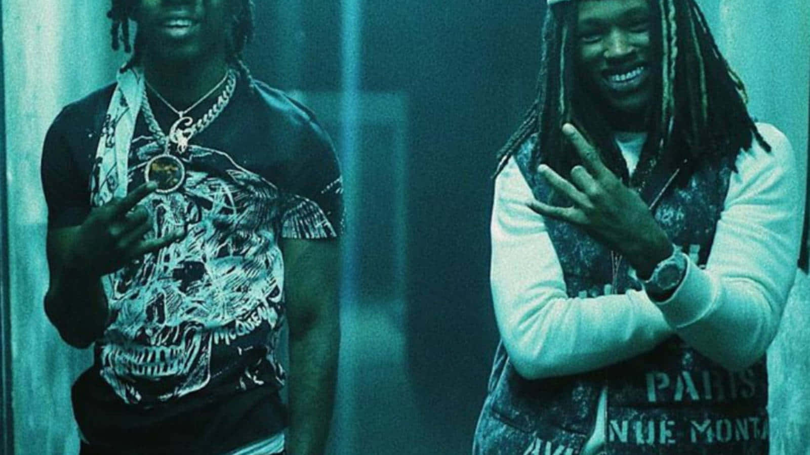 King Von 桌布 壁紙 — 兩位面帶微笑、梳著辮子的男士在昏暗的房間中自信地擺姿勢，房間沐浴在青綠色的燈光下，他們穿著圖案T恤和戴著鏈條。