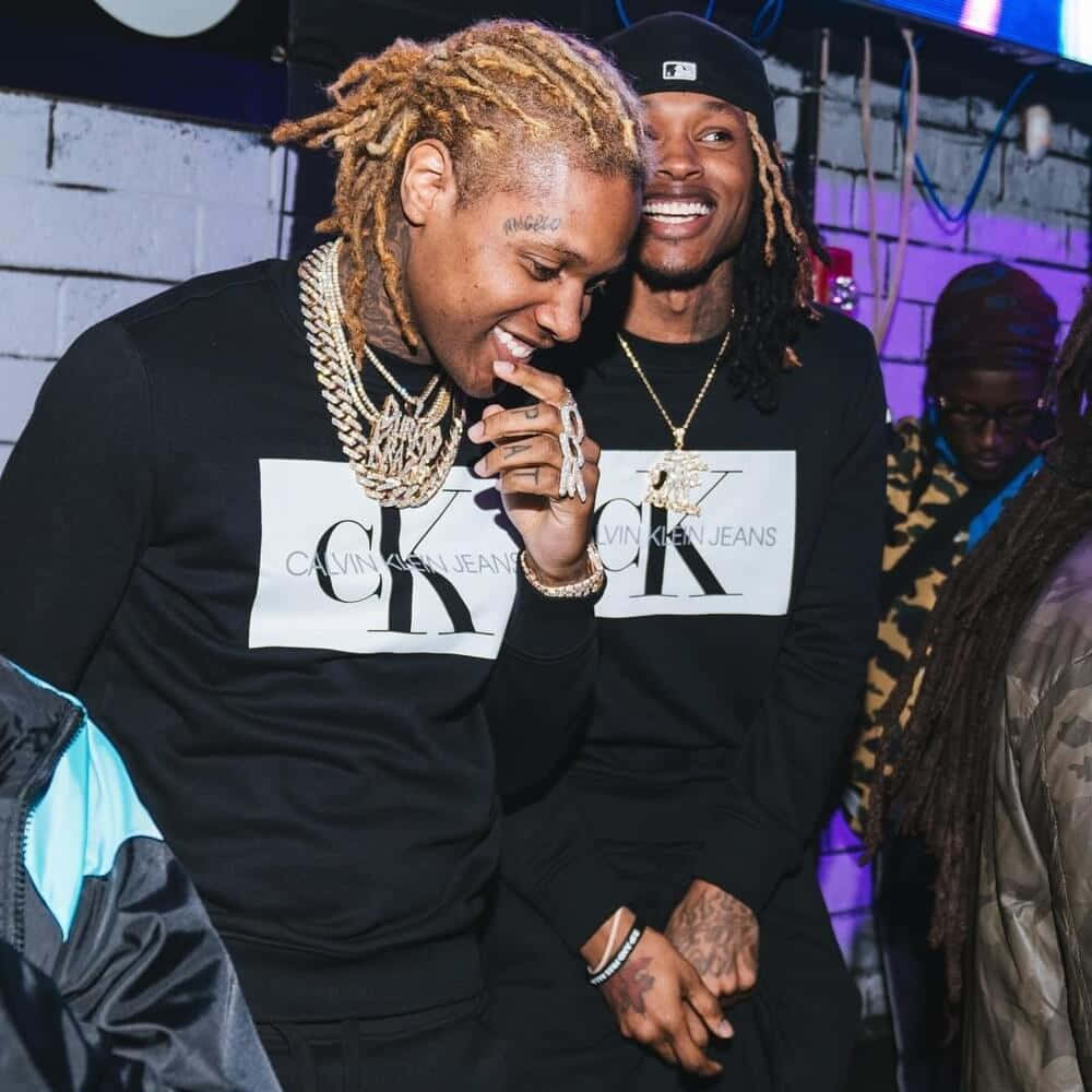 King Von 桌布 壁紙 — 兩位面帶微笑、梳著辮子頭、戴金鏈的男子，在霓虹燈派對上穿著相配的Calvin Klein Jeans運動衫一起大笑。