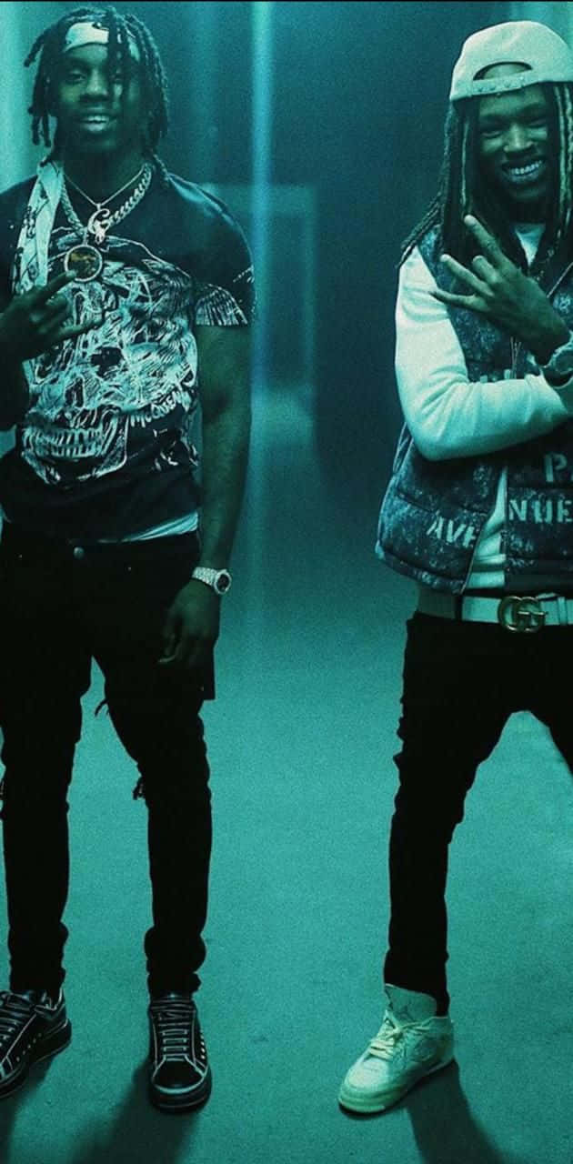 King Von 桌布 壁紙 — 兩位面帶微笑、留著辮子的男士，在光線昏暗的藍綠色房間裡，穿著街頭服飾和配飾，自信地擺姿勢。