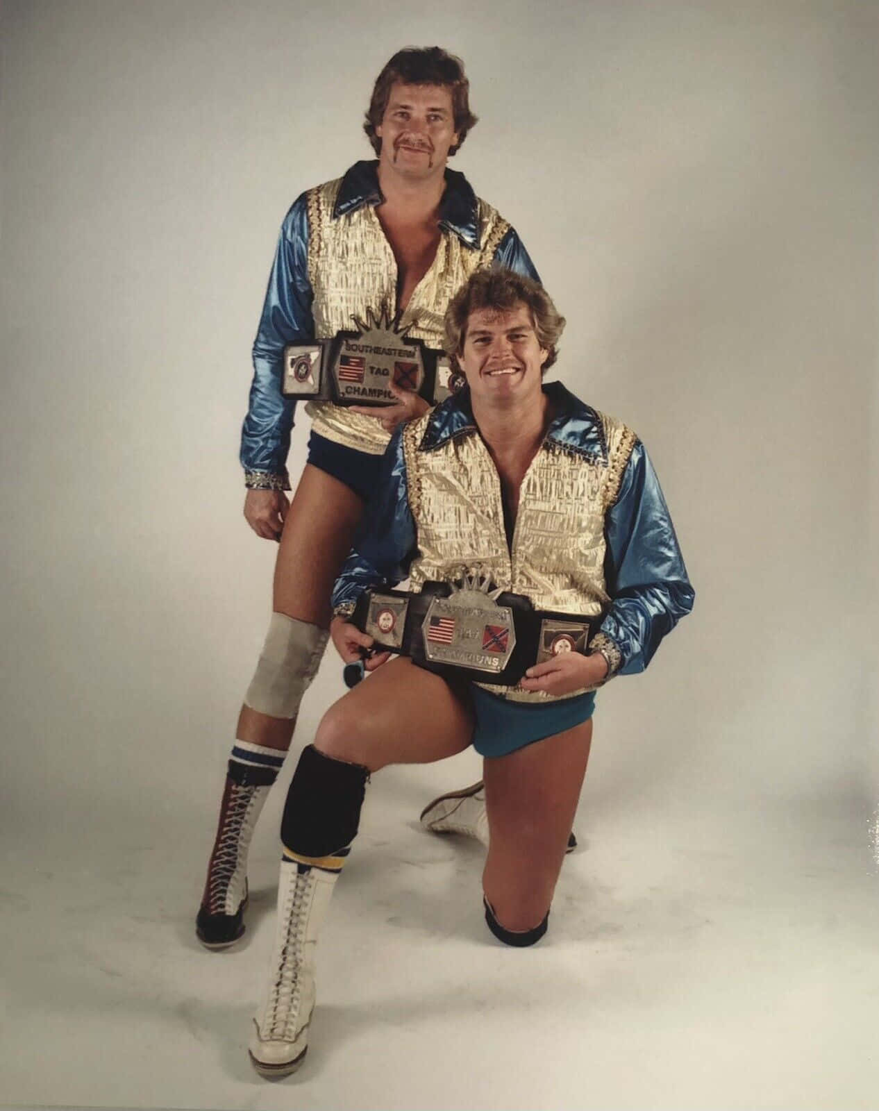Tag Team Wrestlers Robert Fuller And Jimmy Golden Full Shot 壁紙 — 兩位面帶微笑的1980年代摔角手，身穿閃亮藍色和金色外套，在樸素的工作室背景前自豪地展示冠軍腰帶。