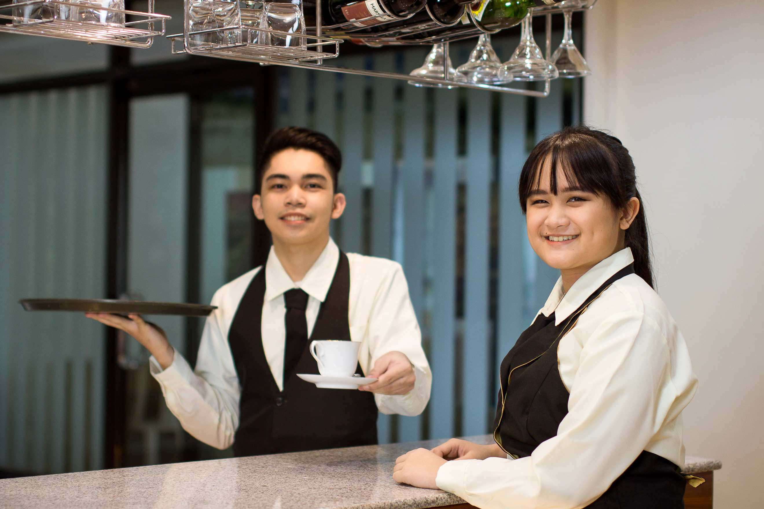 Hospitality 桌布 壁紙 — 兩位面帶微笑的侍應生穿著整潔制服，在現代酒店大堂酒吧提供茶和葡萄酒。