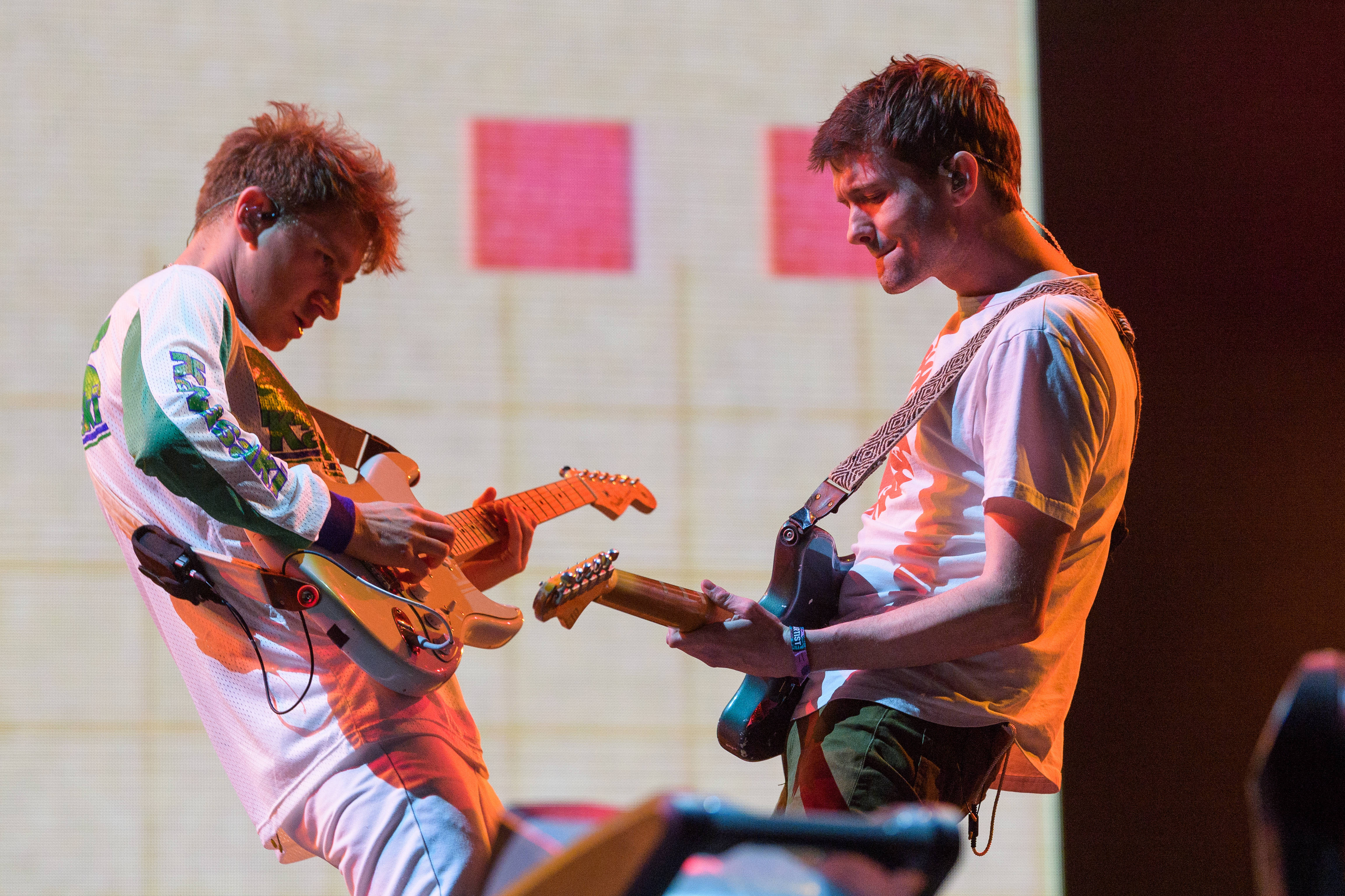 Glass Animals Drew 壁紙 — 兩位音樂家在溫暖的舞台燈光下，於現場音樂會表演中激烈地彈奏電結他。