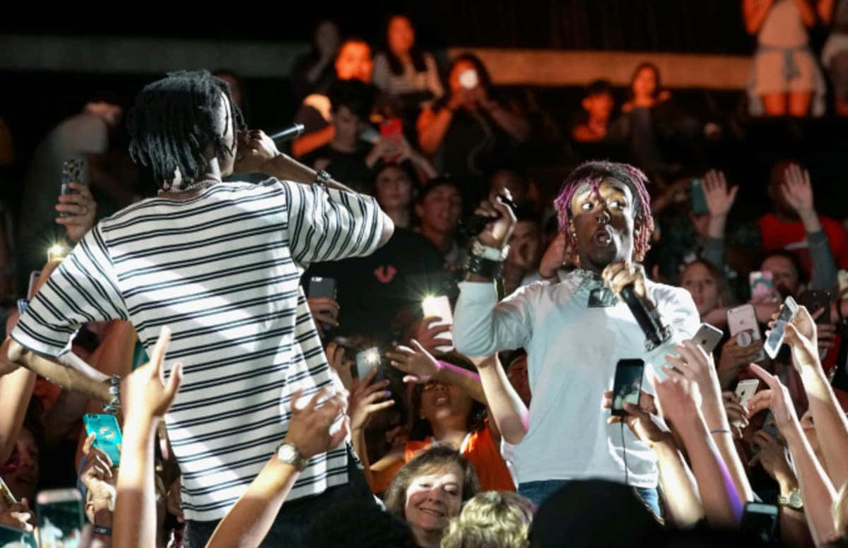 playboi carti 桌布 壁紙 — 兩位饒舌歌手在夜間舞台上充滿活力地表演，周圍是熱情的人群高舉發光手機。