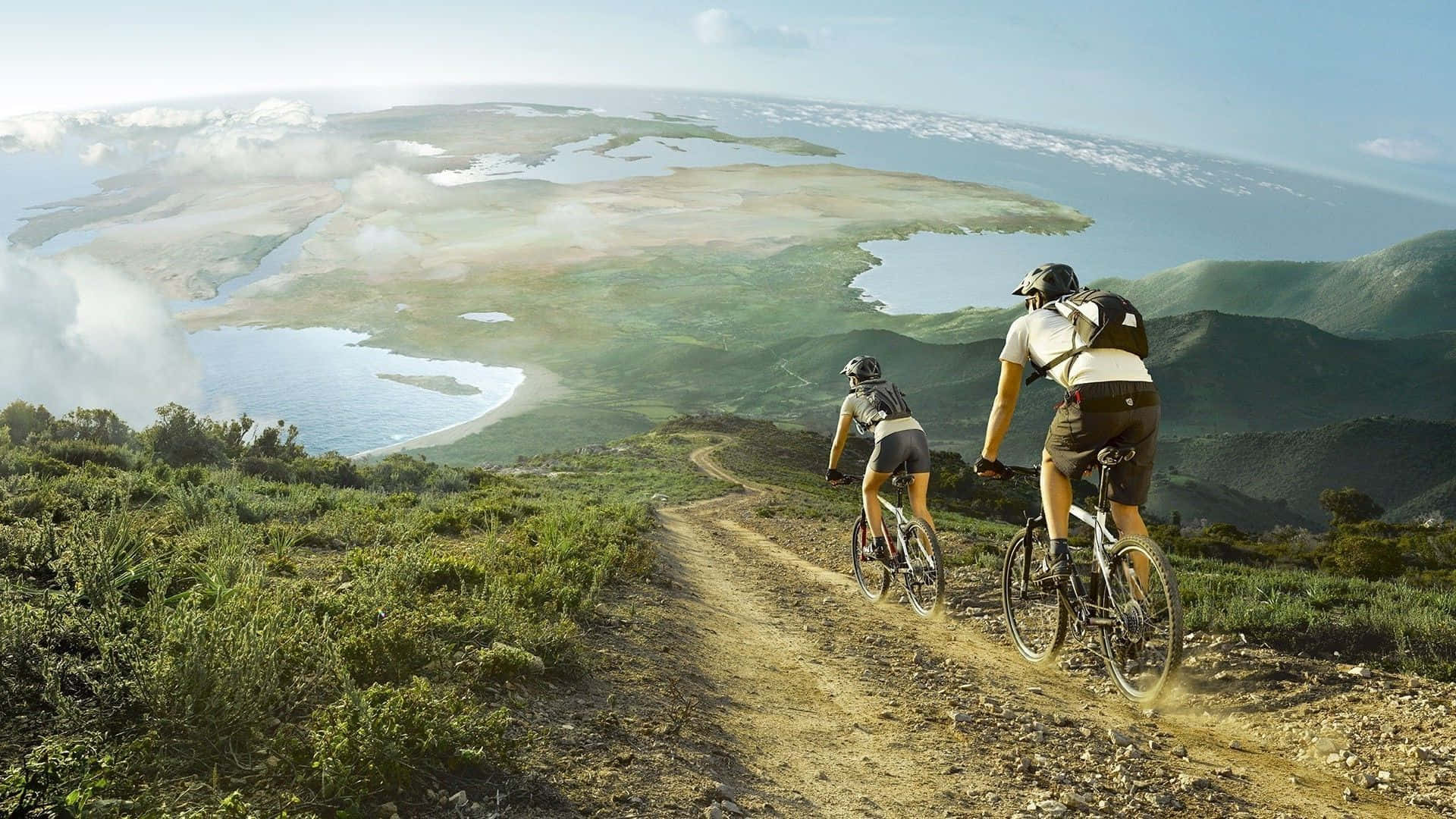 Mtb 桌布 壁紙 — 兩位騎單車人士在泥路上踩踏上山，俯瞰廣闊的沿海景觀，蜿蜒的水道和綠色的山丘在明亮的天空下。