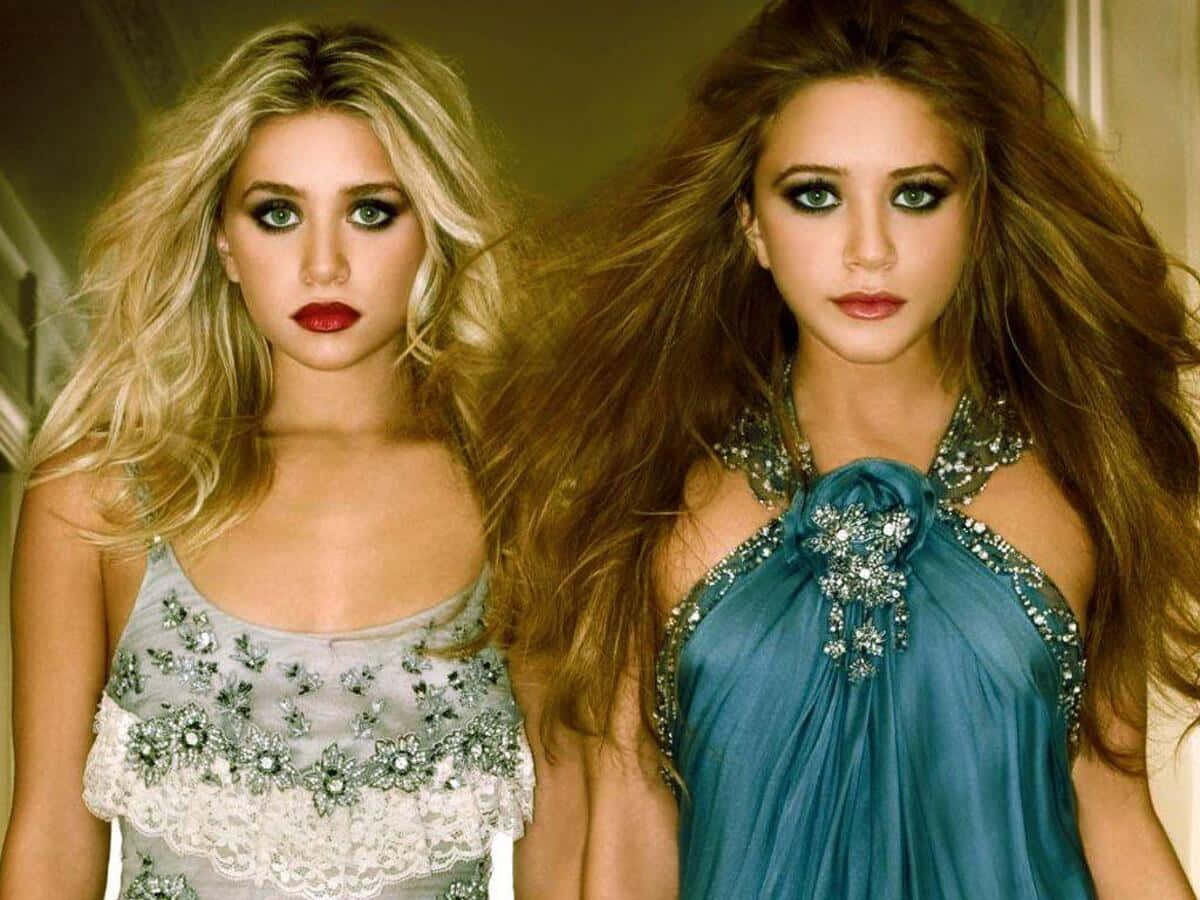 Mary Kate Olsen 桌布 壁紙 — 兩位魅力四射的女性，妝容大膽，秀髮飄逸，在溫暖的攝影棚燈光下穿著華麗的連衣裙擺姿勢。