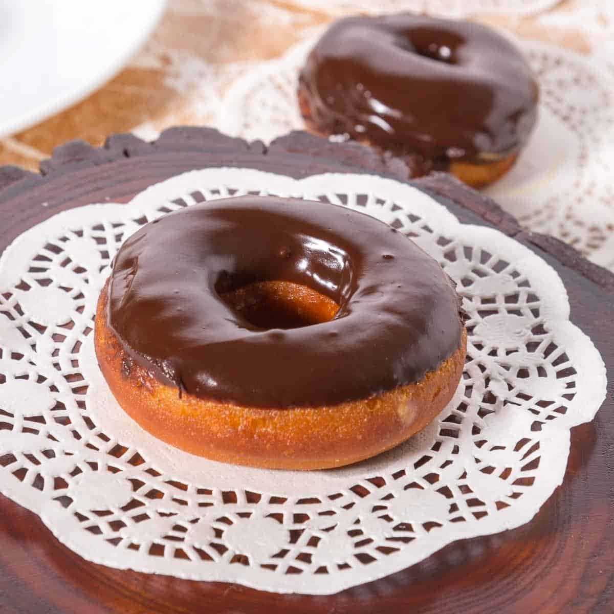 Glazed Donut 桌布 壁紙 — 兩個亮面朱古力糖霜冬甩放在深色木托盤上的蕾絲墊上。