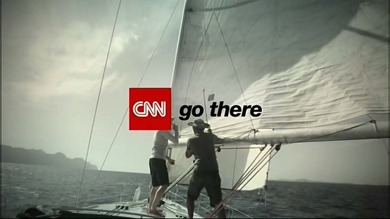 CNN Go There 壁紙 — 兩個人在多雲的海上駕駛一艘揚起白色帆的船，遠處有陸地。